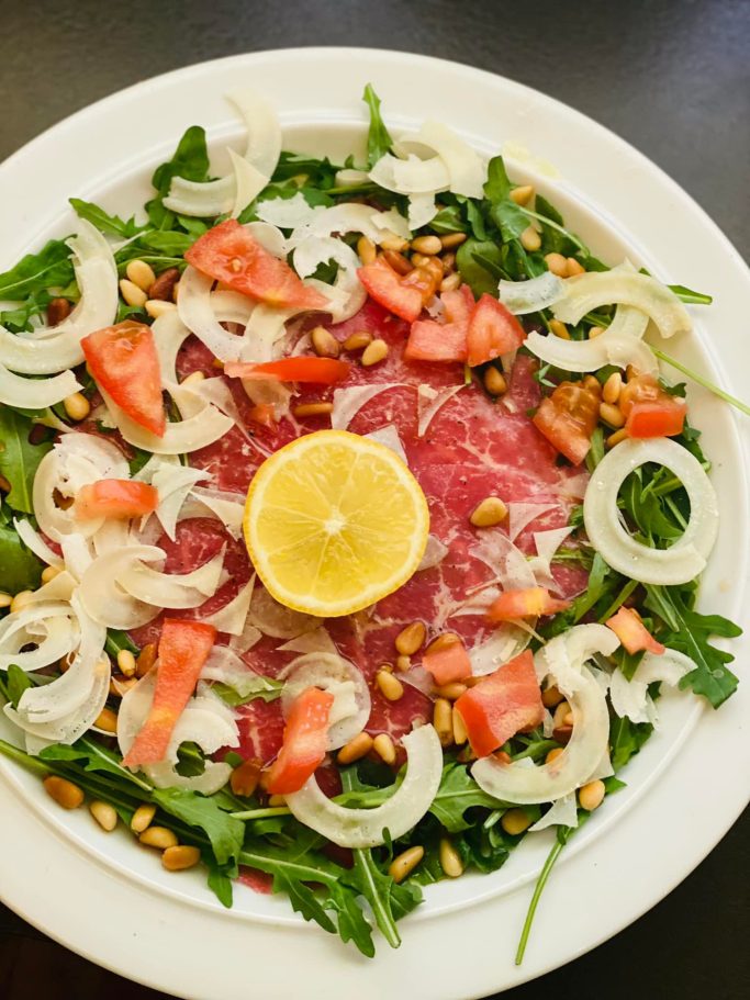 Carpaccio mit Rucola, Tomaten, Zwiebeln, Pistazien und einer Zitronenscheibe.