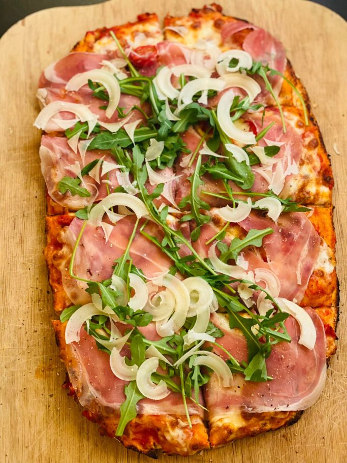 Pinsa mit Schinken, Zwiebeln und Rucola auf einem Holzbrett.