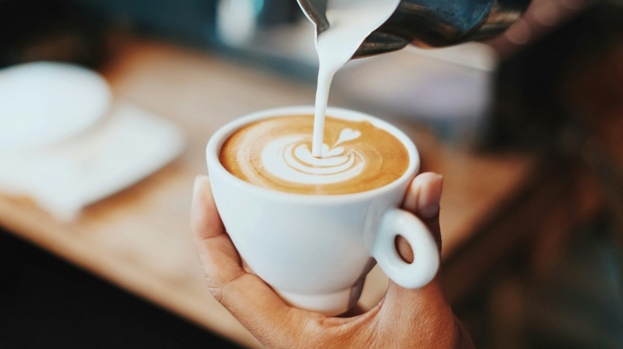 Eine Hand gießt Milch in eine Tasse mit Kaffee, die ein Muster bildet.